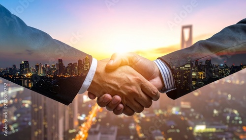 Double exposure photograph of a professional handshake combined with a modern cityscape at sunrise.