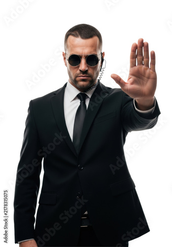 mid-40s caucasian man in tailored black suit and dark sunglasses, security earpiece, raising his hand in a forceful stop gesture on a stark white studio background. concept of security and authority