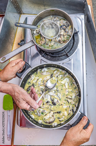 A chef using a spoon to taste and check the pil-pil emulsion sauce of Kokotxas al pil pil, with a small pot simmering cod skins and bones for broth nearby.