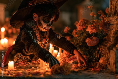 Boy in skeleton costume and skull makeup placing marigolds by glowing candles on a dark granite grave for family remembrance and Day of the Dead