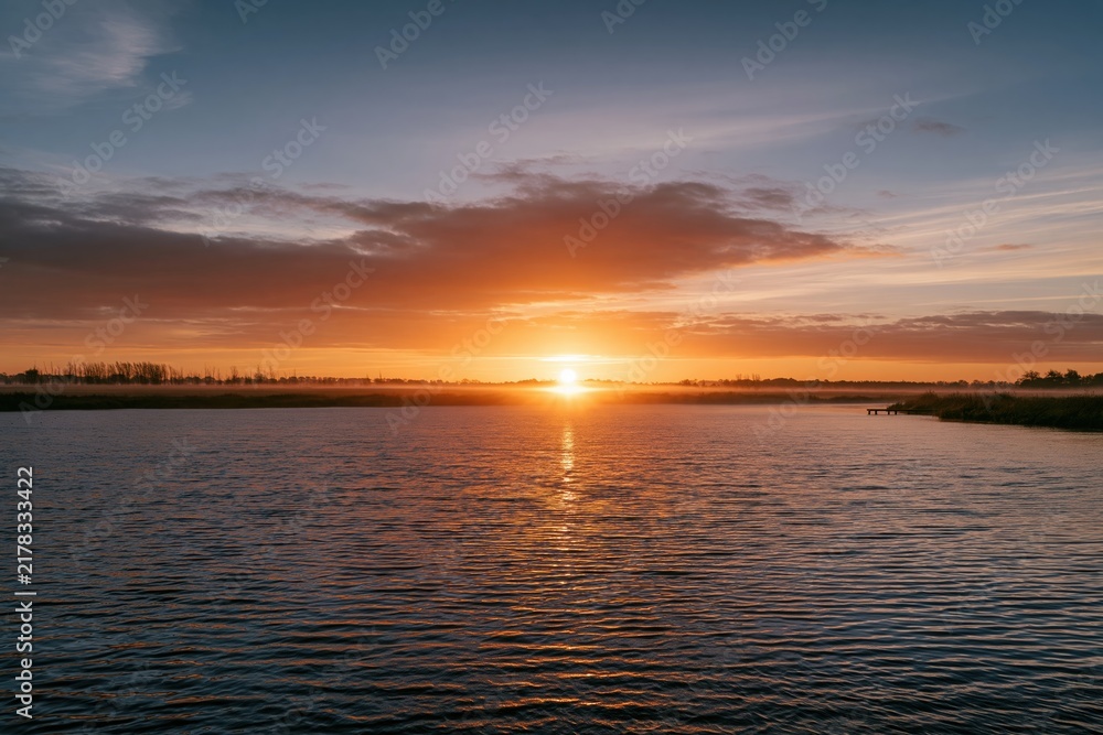 Obraz premium Glowing maple leaf sun over a misty river valley at dusk, featuring deep amber light and calm slate-grey water for autumn travel and seasonal campaigns, with copy space