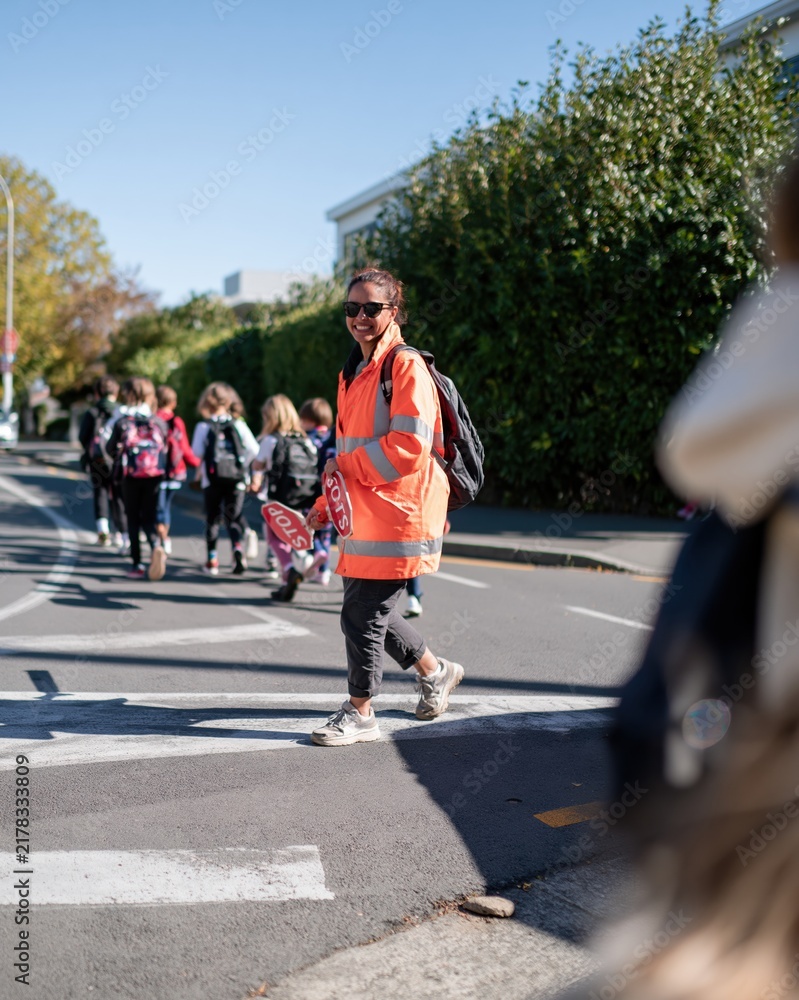 Fototapeta premium Hispanic crossing supervisor in a bright orange hi-vis coat guiding children across a sunny street, with copy space for road safety and back to school programs