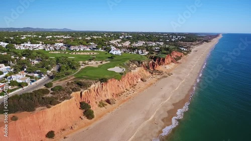 Aerial view flying along the beautiful coastline of vale do lobo, showing the iconic red cliffs, sandy praia de garrao, and a luxury golf course under a clear blue sky in algarve, portugal