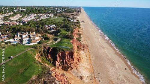 Aerial drone view flying along the stunning coastline of praia de garrao in vale do lobo, portugal, revealing red cliffs, a long sandy beach, and luxurious villas with a golf course