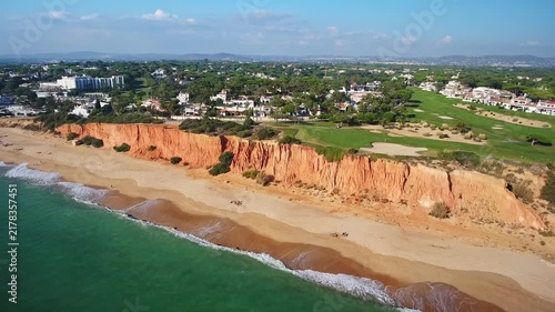 Dramatic aerial footage showing the beautiful red cliffs of garrao beach bordering the green vale do lobo golf course, a popular tourist destination in the algarve region of southern portugal