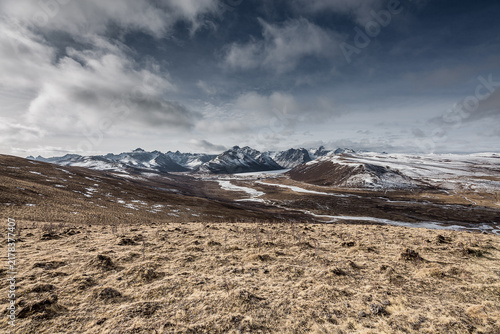 Foto A vast high-altitude landscape featuring snow-capped mountains, dry grassy terrain, and a winding river under a cloudy sky