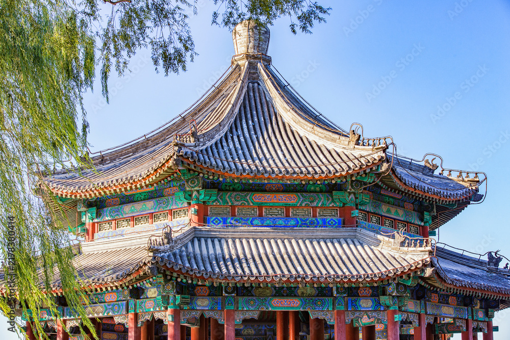 Fototapeta premium A two-tiered traditional Chinese pavilion with ornate painted decorations and curved tile roofs, set outdoors under a clear blue sky with willow branches nearby.