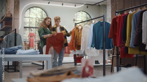 Clothing Store Shopping: Young Caucasian Woman Searches a Rail of Colorful Apparel Before Her Boyfriend Joins Her, Sharing Opinions and Laughing Together During a Casual Fashion Shopping Trip.