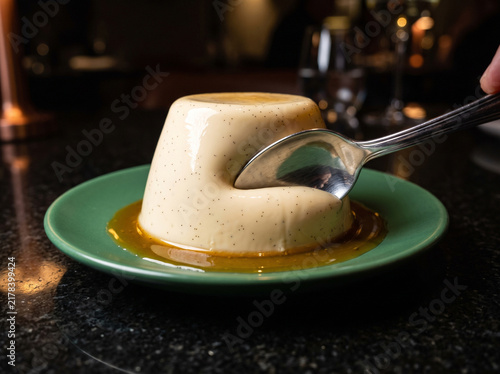 Spoon pressing into a soft vanilla panna cotta dessert with caramel sauce on a green plate