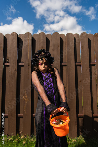 Quadro su tela Little girl in stylish Halloween costume holding a bucket for treats against a w