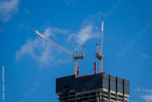 Construction cranes work on a tall building under a blue sky with some clouds in the background during the day