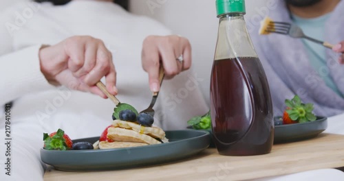 Two adults are pressing fork and cutting blueberry pancakes on wooden lap tray to share breakfast