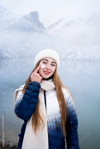 Obraz na plátně Young woman applying moisturizing face cream while standing beside a snowy mountain lake, representing winter skincare, skin hydration and protection from cold weather