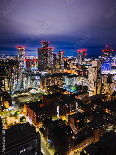 Manchester UK: 16th August 2026: A drone view of the Manchester skyline near Central shows buildings illuminated by city lights at night. Construction activity is visible