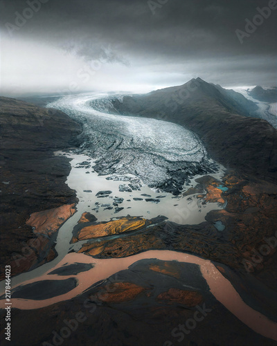 Fotografía Massive glacier flows through a dark mountain valley into braided meltwater streams, creating a dramatic aerial landscape ideal for climate, geology, travel and environmental content