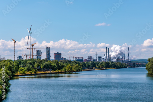 Obraz na plátne Industrial complex with smokestacks emitting steam and wind turbine along waterw