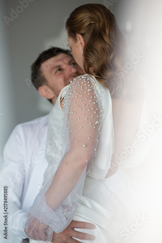 Bride in white wedding dress leaning over groom on bed during calm wedding morning. Intimate moment, soft light, emotional connection, modern wedding lifestyle indoors.
