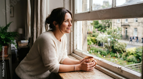 Woman holding a mug looking out a window at a garden and buildings