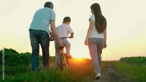Parents teach child to ride bicycle in park. Happy family. Young family with little son riding bicycle. Family with child boy playing at sunset. Teamwork, dad mom son. Sport family. Father mother son