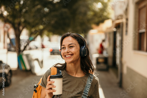 Young woman smiling walking city street with headphones and coffee