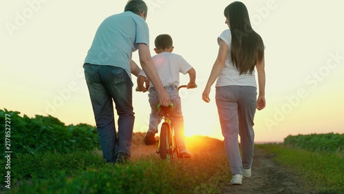 Parents teach child to ride bicycle in park. Happy family. Young family with little son riding bicycle. Family with child boy playing at sunset. Teamwork, dad mom son. Sport family. Father mother son