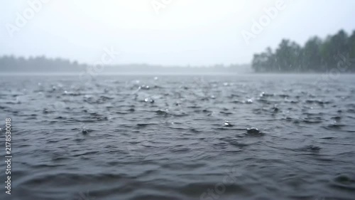 Raindrops falling on a lake surface creating ripples and splashes with a misty forest background in slow motion footage with against blurred background background trees distance