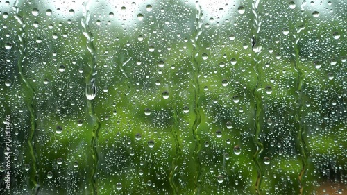 Raindrops sliding down a glass window pane with a blurred green forest in the background with trickling down transparent water droplets trickling surface during rainstorm