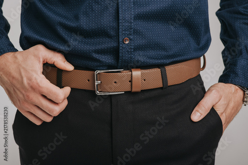 Close-up of a man wearing a light brown perforated leather belt with a classic metal buckle on black trousers