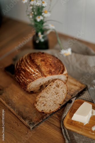 Freshly baked artisan bread sits warm on wooden board with butter and wildflowers nearby