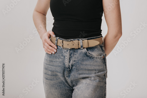 Close-up of a woman wearing a light beige smooth leather belt with a classic metal buckle on acid wash jeans