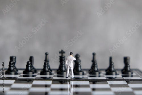 Obraz na płótnie Miniature businessman standing on chessboard facing opponent pieces, business st