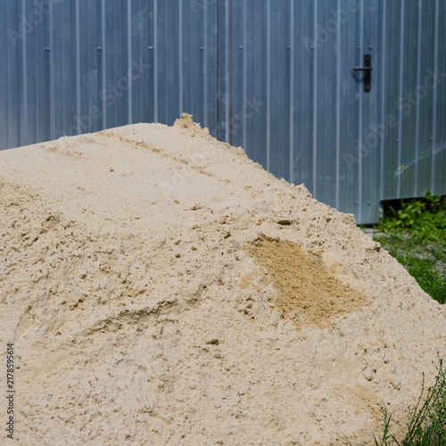 A big pile of yellow construction sand lying on the ground next to a corrugated metal fence.