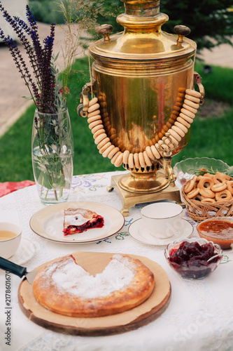 Bild auf Leinwand A golden samovar with bagels, tea cups, homemade pie, and jam on a white tablecloth set for tea in a garden