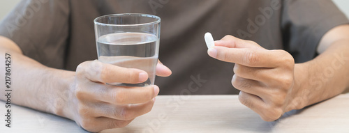 Bild auf Leinwand Sick asian young man holding tablet pill on hand, painkiller medicine from fever, stomach pain, headache for treatment, take drug, vitamin with glass of water on table at home