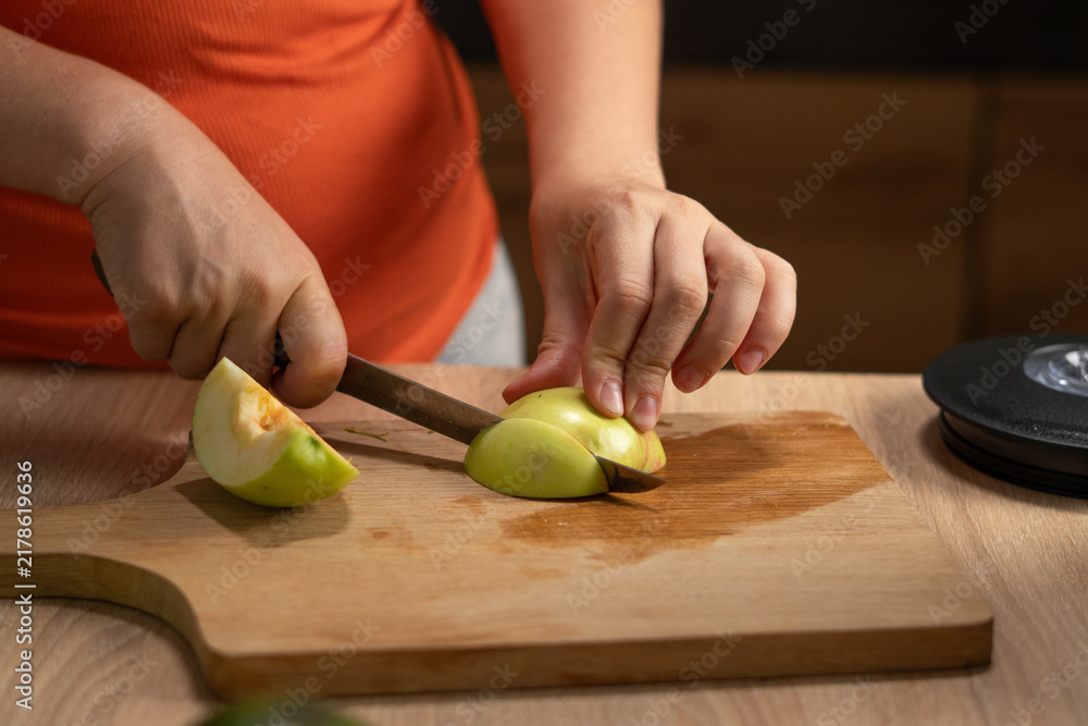 Obraz premium Woman slices a fresh green apple on a wooden cutting board in the kitchen, with her hands holding the fruit and knife.