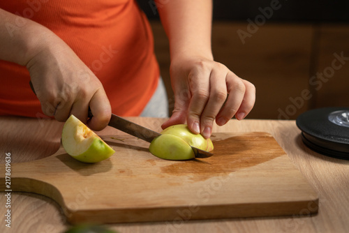 Woman slices a fresh green apple on a wooden cutting board in the kitchen, with her hands holding the fruit and knife.