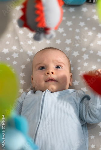 Cute infant baby lying in crib and looking up at hanging colorful mobile toys in a cozy nursery.
