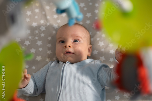 Cute infant baby lying in crib and looking up at hanging colorful mobile toys in a cozy nursery.