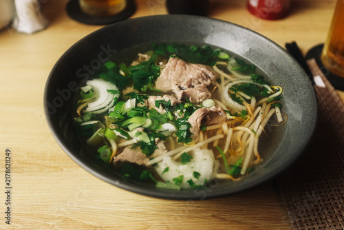 Traditional Asian noodle soup with sliced beef bean sprouts onion and fresh herbs served in a dark bowl. Hot clear broth and noodles create a comforting restaurant food background.