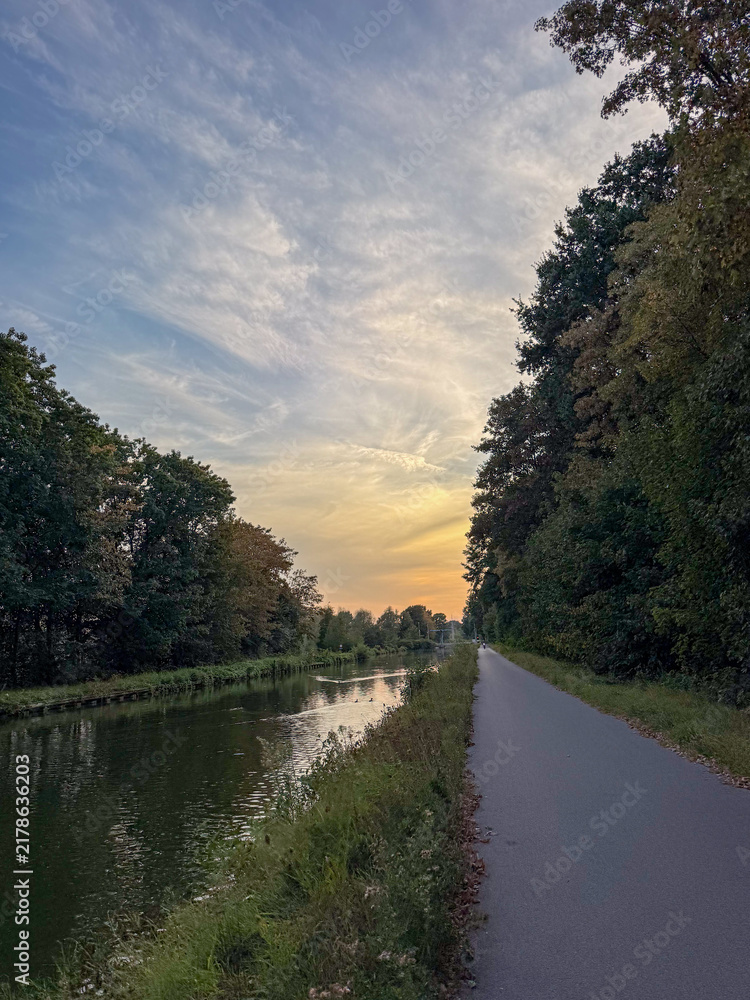 Fototapeta premium Tranquil Canal Towpath at Sunset with Tree-Lined Waterway and Dramatic Golden Sky
