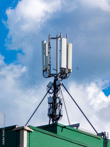Communication antenna mounted on a rooftop against a partly cloudy sky. The tower consists of multiple white rectangular panels and various electronic components attached to a metal frame. Stabilizing