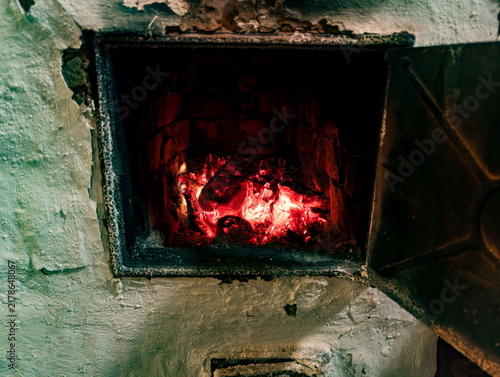Rustic fireplace with glowing red embers providing warm, ambient light. The hearth shows signs of use with soot-stained bricks and surrounding textured, weathered plaster walls. Metal door slightly aj