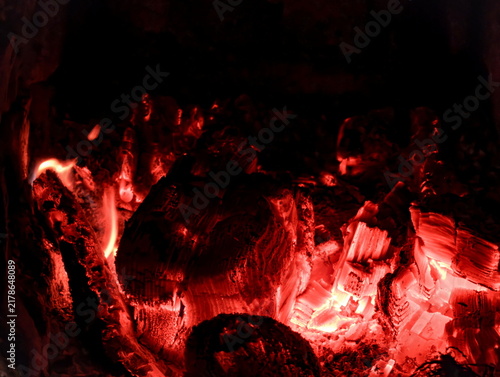 Close-up view of glowing embers producing intense red and orange light, highlighting the texture of burnt wood and ashes. Dark background contrasts with the bright molten coals, showcasing heat and en