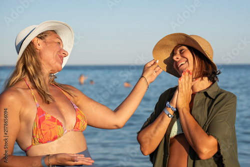 Fotografía Two generation x women laugh and playfully adjust a sun hat while enjoying a carefree summer day at the beach