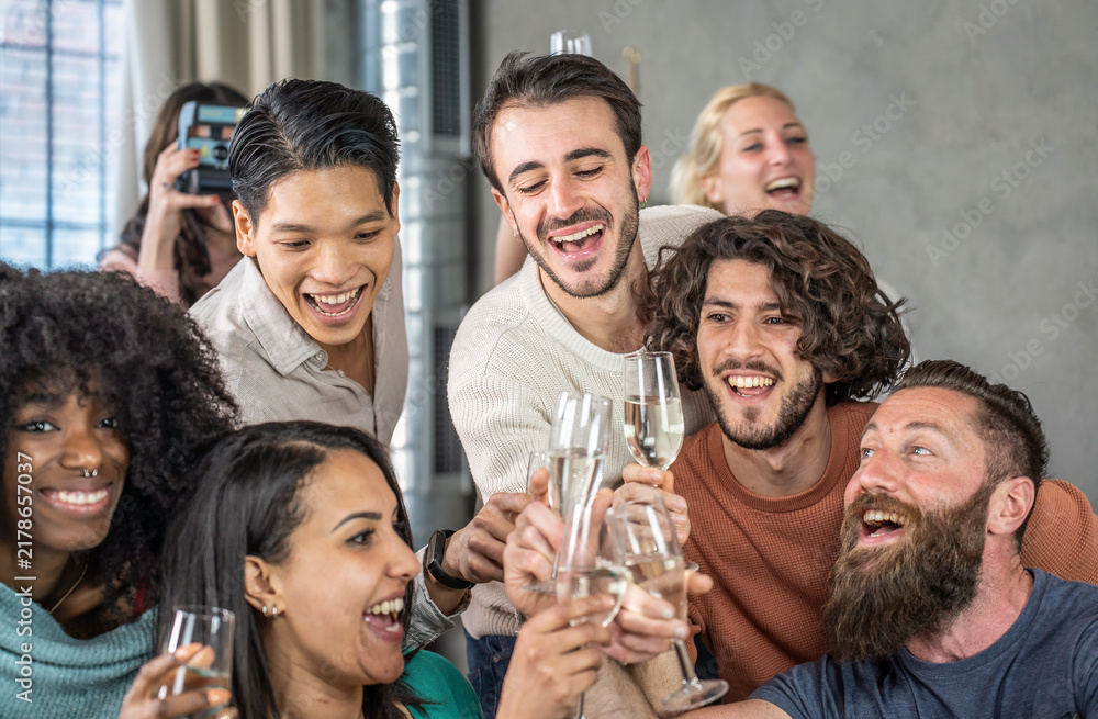 Obraz premium Joyful multiethnic friends clink sparkling wine glasses in a close-up group toast during a lively celebration at home. Smiling young adults share laughter, friendship, and togetherness.