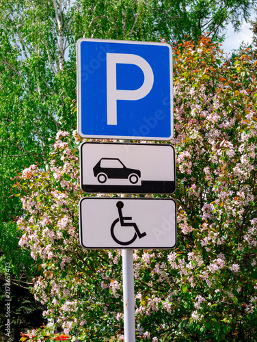 Blue parking sign mounted above two white panels, one depicting a car icon and the other showing a wheelchair symbol, indicating designated parking space for disabled vehicles. Lush greenery and flowe