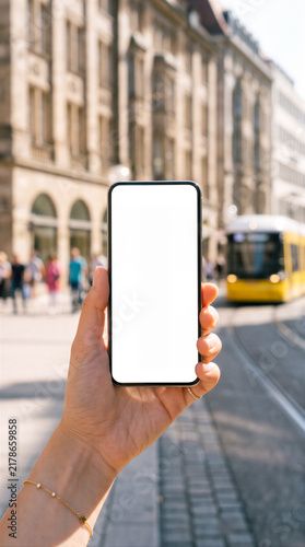 Close-up of a hand holding a smartphone with a blank white screen mockup against a blurred sunny city street with a yellow tram