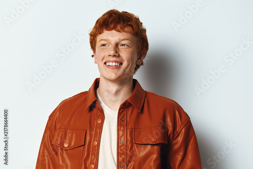Konstfotografi Young man with red hair and smiling face posing in brown leather jacket on light background for fashion and portrait photography