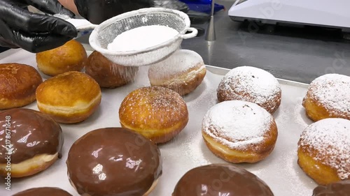 Baker in black gloves sprinkles powdered sugar on fresh donuts through sieve. Chocolate glazed doughnuts on baking tray. Pastry preparation and bakery production concept