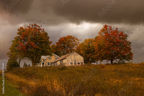 Historic wooden barn and farmhouse surrounded by colorful autumn trees under storm clouds, Ontario, Canada.